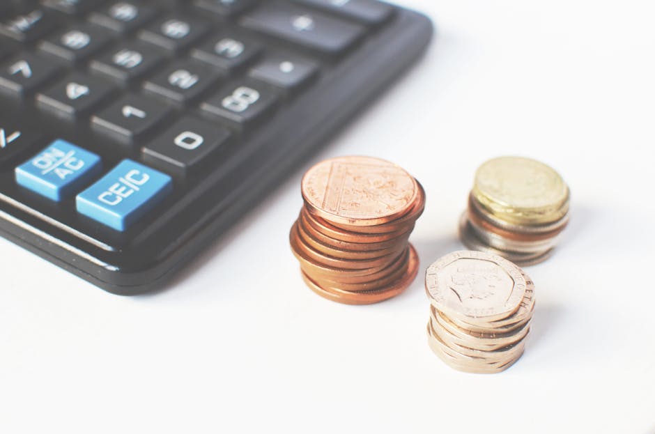 Close-up image of a black calculator with light blue buttons positioned on the left side of the frame, accompanied by three stacks of Euro coins—one copper-colored stack of five-cent coins, a small stack of gold-colored one-euro coins, and a taller stack of mixed denomination coins—placed on a white background. The coins are arranged in separate piles, with some slightly overlapping, illustrating financial planning or cost estimation potentially related to house removals or relocation costs. The image emphasizes the connection between budgeting and moving services, with the presence of the calculator and coins symbolizing the financial considerations involved in home relocation, packing, and furniture transport processes that Man With a Van Leyton might assist with, especially when dealing with average prices and hidden costs highlighted in the page about Leyton removals prices.