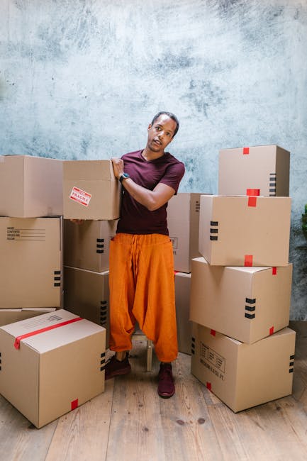 A person with medium skin tone, wearing a maroon t-shirt and bright orange trousers, stands indoors surrounded by stacked cardboard moving boxes with red and black tape, some with printed shipping labels. The individual is holding a box with one hand and resting the other on top of another box, standing on a wooden floor against a textured light blue wall. Visible on the left are additional boxes placed on the floor, some in the process of being packed or organized for a home relocation. The scene depicts the loading process involved in furniture transport or packing and moving, reflecting the services offered by Man With a Van Leyton. There is natural ambient lighting illuminating the space, highlighting the preparation and logistics involved in removals within the context of house moves or moving logistics.