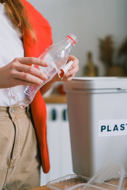 A person wearing a white shirt and beige trousers, with a red cardigan partially visible, is holding a clear plastic bottle over a white recycling bin labeled 'PLASTIC' in black letters. The individual is in the process of disposing of a used plastic bottle, which is seen to be partially crushed with red and white cap. The scene takes place indoors near a kitchen or utility area, with a wooden countertop and other containers or items in the background. The lighting is soft and natural, emphasizing the act of sorting recyclable waste as part of home relocation or packing and moving activities. The image reflects the importance of proper waste separation during house removals, and the focus on environmentally responsible disposal aligns with the services provided by Man With a Van Leyton in Lewton, supporting efficient and sustainable furniture transport and packing processes.