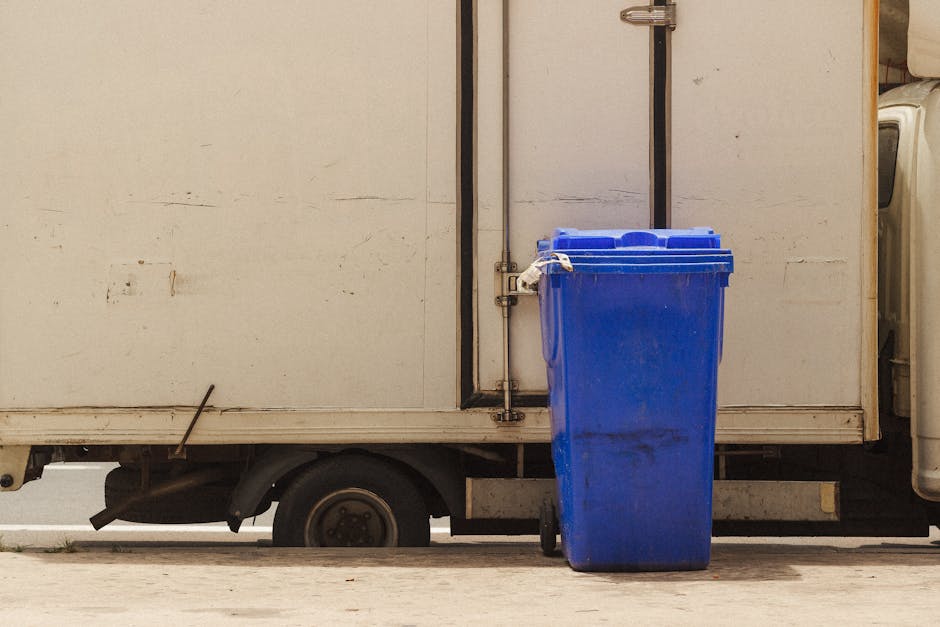 A white delivery truck with a partially visible sliding side door parked outdoors on a paved surface. Attached to the side of the truck is a large blue recycling bin standing upright, positioned on the pavement near the side door. The bin has a closed lid and appears to be made of plastic. The image captures the lower section of the truck, including its black wheel and part of the undercarriage, with the background showing a neutral outdoor setting. The scene may be related to home relocation or moving activities, and Man With a Van Leyton's removal services are associated with managing waste disposal or logistics during moves.