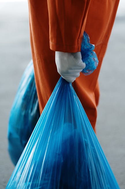 A person wearing a white shirt and beige trousers, with a red cardigan partially visible, is holding a clear plastic bottle over a white recycling bin labeled 'PLASTIC' in black letters. The individual is in the process of disposing of a used plastic bottle, which is seen to be partially crushed with red and white cap. The scene takes place indoors near a kitchen or utility area, with a wooden countertop and other containers or items in the background. The lighting is soft and natural, emphasizing the act of sorting recyclable waste as part of home relocation or packing and moving activities. The image reflects the importance of proper waste separation during house removals, and the focus on environmentally responsible disposal aligns with the services provided by Man With a Van Leyton in Lewton, supporting efficient and sustainable furniture transport and packing processes.