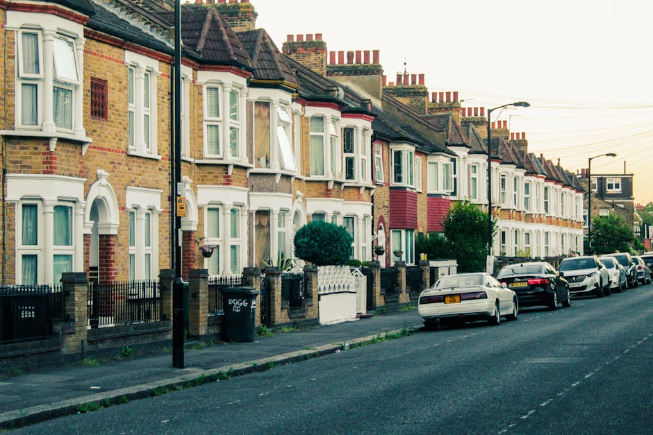 A row of Victorian terraced houses on Francis Road in Leyton, featuring characteristic bay windows with white frames and brick facades in various shades of red, brown, and cream. Some houses have front gardens enclosed by low brick walls with black metal railings, and potted plants or small shrubs are visible in a few. Parked cars, including sedans and compact vehicles, line the street directly in front of the homes, with a driveway partly visible on the left. A lamppost stands on the pavement, which is made of concrete slabs, and the street scene is illuminated by soft, natural light, suggesting late afternoon or early evening. The environment reflects a residential area suitable for house removals, containing elements like parking, street lighting, and typical residential architecture.