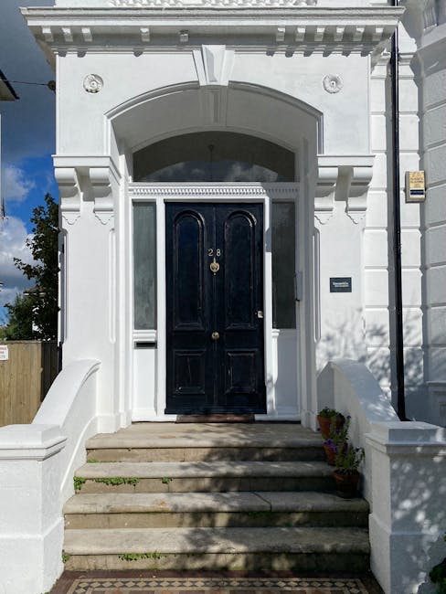 The image shows the front entrance of a narrow Victorian terraced house with a white facade and decorative architectural details above the doorway. The dark-colored front door, with the house number 28 displayed in gold, is centrally positioned and accessed via a concrete staircase with five steps. To the right of the door, there is a small black mailbox mounted on the wall, and on the left, a short white wall extends from the building, forming a small boundary. The entrance area is shaded by shadows of nearby trees, and a few potted plants are placed at the base of the staircase. The surrounding environment includes a wooden fence visible on the left side, and the sky above is partly cloudy with patches of blue. This exterior setting reflects a typical residential property involved in home relocation and furniture transport, as part of professional removals, such as those by Man With a Van Leyton, supporting house removals and packing for narrow Victorian homes like the one on Francis Road.