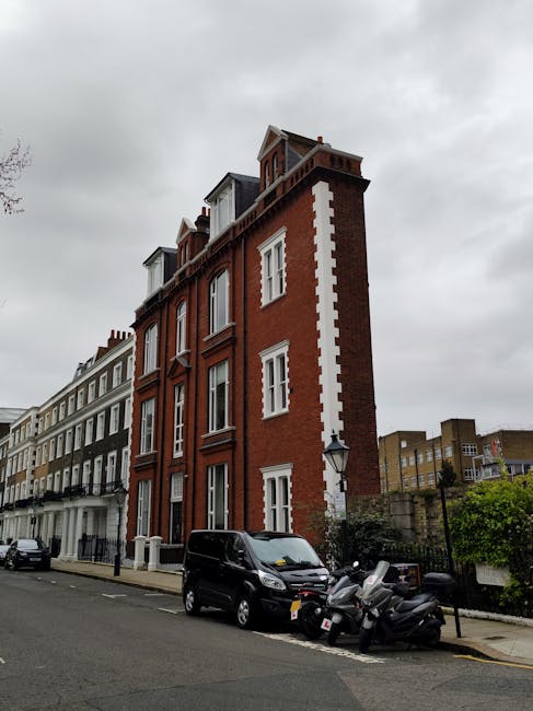 A row of Victorian terraced houses on Francis Road in Leyton, featuring characteristic bay windows with white frames and brick facades in various shades of red, brown, and cream. Some houses have front gardens enclosed by low brick walls with black metal railings, and potted plants or small shrubs are visible in a few. Parked cars, including sedans and compact vehicles, line the street directly in front of the homes, with a driveway partly visible on the left. A lamppost stands on the pavement, which is made of concrete slabs, and the street scene is illuminated by soft, natural light, suggesting late afternoon or early evening. The environment reflects a residential area suitable for house removals, containing elements like parking, street lighting, and typical residential architecture.