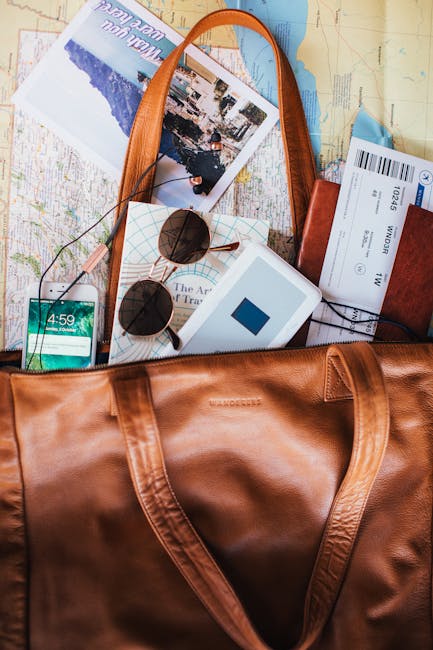 A collection of travel and packing items spread out on a surface, including a large brown leather duffle bag with open zipper, a pair of black sunglasses with round lenses resting on a small white box, a smartphone displaying the time 4:59, a camera or portable device partially visible, a map or travel guide with a photograph of a coastal scene, an airline boarding pass, a brown leather wallet, and a pair of wired earphones. The background features a light-colored wooden surface, indicating a packing or home relocation process. These objects are associated with house removals, packing, and preparation for transport, aligned with services offered by Man With a Van Leyton for Leyton High Road flats.