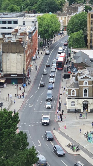 An aerial view of Leyton High Road with a line of vehicles, including a red bus, cars, and vans, traveling along the street surrounded by buildings, including some with brick facades and others with modern design, as well as green trees lining the pavement. Pedestrians are visible walking on the sidewalks, and a few bicycles are parked near a small building at the corner. The scene captures daytime urban moving activity, suggesting ongoing home relocations or furniture transport processes in the area, which aligns with local house removals services such as those offered by Man With a Van Leyton. The street appears busy but orderly, with clear road markings and traffic signals, emphasizing the importance of planning for efficient moving logistics within the Leyton E10 area.