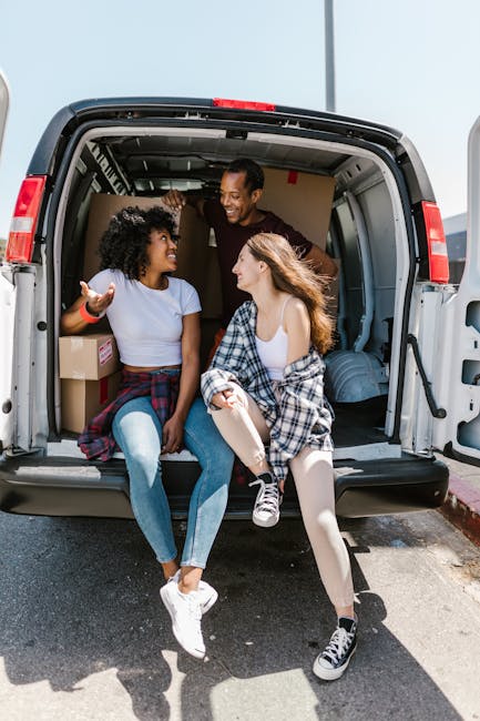 Two young women sit on the edge of an open van's loading area during a home relocation, engaging in a conversation and smiling at each other. One woman, with curly black hair, wears a light blue t-shirt, blue jeans, and white sneakers, while gesturing with her hand. The other woman, with long red hair, wears a white tank top under a flannel shirt tied around her waist, light-colored tights, and black and white sneakers. Inside the van, moving boxes made of cardboard are visible, some wrapped with plastic for protection, along with packing materials and furniture items. The van, which is parked on a residential street, is equipped with a ramp or lift for loading and unloading, and is under natural daylight, indicating daytime. This scene depicts the packing and moving process, highlighting the involvement of Man With a Van Leyton in furniture transport and local removal services, with a focus on organized, efficient home relocation logistics.