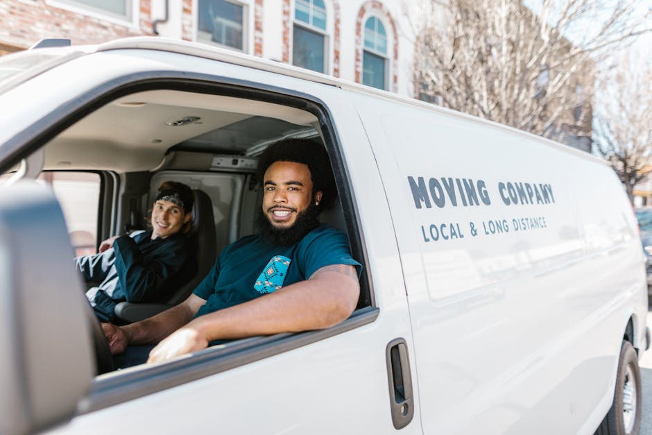 Inside the cab of a white moving van belonging to Man With a Van Leyton, two movers are seated and smiling at the camera. The driver, a man with dark curly hair and a beard, is positioned in the driver's seat, wearing a dark blue T-shirt with service branding. Next to him, a woman with dark hair pulled back is seated in the passenger seat, also smiling and dressed in casual clothing suitable for a home relocation crew. The van's interior features equipment for furniture transport and packing, with visible straps and blankets stored inside. The exterior of the van displays the company's name and services, including 'LOCAL & LONG DISTANCE' removals, and is parked in an urban setting near Leyton Jubilee Park. The background shows building facades and leafless trees, indicating a typical local area in Leyton, where the company provides packing and moving services, including loading and transportation for house removals, often involving careful handling of boxes, furniture, and appliances.