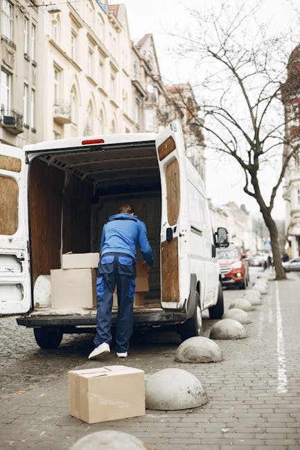 A man wearing a blue jacket and dark trousers is seen from behind, loading a large, open white moving van parked on a city street. The van's interior is lined with wooden paneling, and several cardboard boxes are visible inside. One cardboard box is on the pavement directly in front of the van, while another is being handled by the man. The street features lined curb stones and parked cars, with multi-storey, cream-coloured buildings with decorative facades and balconies on the adjacent side. A leafless tree stands across the street, indicating a late autumn or winter season. This scene captures the loading process during a home relocation or furniture transport activity, emphasizing careful handling of packed boxes and household items, with natural daylight illuminating the environment. The image illustrates the logistical steps involved in professional removals, exemplifying the services of Man With a Van Leyton.