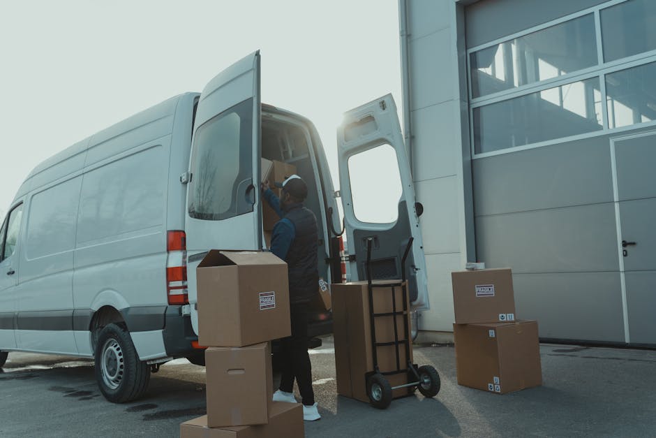 A man wearing a dark jacket and cap is loading cardboard boxes into the rear of a white panel van parked outside a modern building with large glass windows. The boxes are stacked nearby on the pavement, some on a small hand truck and others directly on the ground. The open van door reveals additional packing supplies and possibly furniture or household items inside, indicating a home relocation or moving process. Bright natural light suggests daytime, with the sunlight casting a glow on the van and the loading area. The scene depicts logistical steps in furniture transport and packing during a professional removals service by Man With a Van Leyton, illustrating typical loading procedures associated with residential moves in the LEYTON area. Relevant elements include cardboard boxes, packing equipment, the loading area next to a building, and the van used for furniture transport and home relocation tasks.