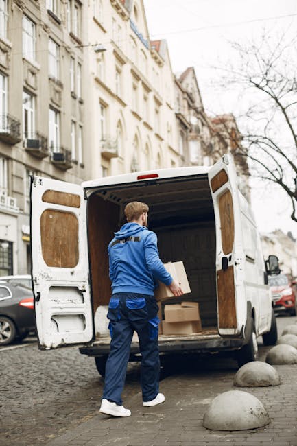 A man wearing a blue jacket and dark trousers is seen from behind, loading a large, open white moving van parked on a city street. The van's interior is lined with wooden paneling, and several cardboard boxes are visible inside. One cardboard box is on the pavement directly in front of the van, while another is being handled by the man. The street features lined curb stones and parked cars, with multi-storey, cream-coloured buildings with decorative facades and balconies on the adjacent side. A leafless tree stands across the street, indicating a late autumn or winter season. This scene captures the loading process during a home relocation or furniture transport activity, emphasizing careful handling of packed boxes and household items, with natural daylight illuminating the environment. The image illustrates the logistical steps involved in professional removals, exemplifying the services of Man With a Van Leyton.
