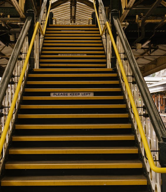 A set of indoor industrial stairs with black and yellow striped treads, flanked by yellow handrails on both sides, leading upward to an overhead platform within a warehouse or station environment. The stairs are made of metal and have a non-slip surface, with a white sign reading 'PLEASE KEEP LEFT' attached to one of the steps, indicating directional instructions for the movement of staff or customers. The surrounding structure includes metal beams, pipes, and industrial lighting, suggesting a commercial or transport setting suitable for a home relocation or furniture transport process, as displays by Man With a Van Leyton often involve careful navigation of such environments during loading or unloading stages.
