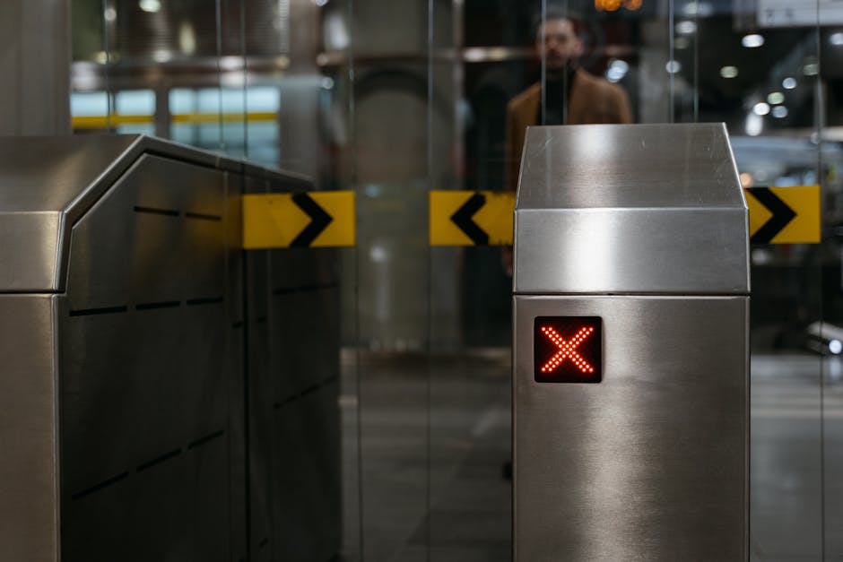 A stainless steel pass-through barrier with a digital display showing a red 'X' in an indoor public transportation or station environment. Behind the barrier, a glass door with black and yellow directional chevrons is visible, indicating restricted access or entry control. The background reflects a well-lit area with faint outlines of people, likely travelers or staff, and the setting appears to be a modern station with metallic fixtures. This scene relates to the logistics of house removals or home relocations, illustrating access points or entry controls often encountered during the moving process, particularly when navigating through stations or transport hubs with a focus on efficient furniture transport and packing logistics.