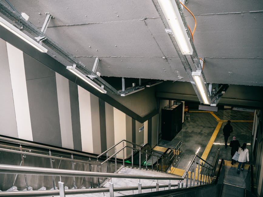 A set of indoor industrial stairs with black and yellow striped treads, flanked by yellow handrails on both sides, leading upward to an overhead platform within a warehouse or station environment. The stairs are made of metal and have a non-slip surface, with a white sign reading 'PLEASE KEEP LEFT' attached to one of the steps, indicating directional instructions for the movement of staff or customers. The surrounding structure includes metal beams, pipes, and industrial lighting, suggesting a commercial or transport setting suitable for a home relocation or furniture transport process, as displays by Man With a Van Leyton often involve careful navigation of such environments during loading or unloading stages.