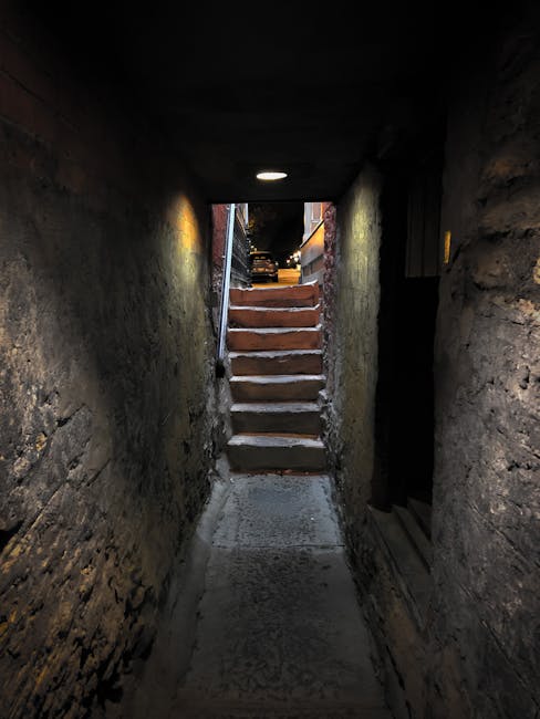 A narrow stairwell leading from an underground passage to an outdoor area during dusk, with wooden steps and a metal handrail on the left side. The corridor is enclosed by stone walls with a rough texture, and the ceiling has a small circular light illuminating the pathway. At the top of the stairs, a black van is partially visible parked outside on a pavement, suggesting the area is used for loading or unloading. The setting indicates a home relocation or furniture transport process, with visible cardboard boxes and moving equipment possibly outside, supporting the context of house removals services by Man With a Van Leyton.