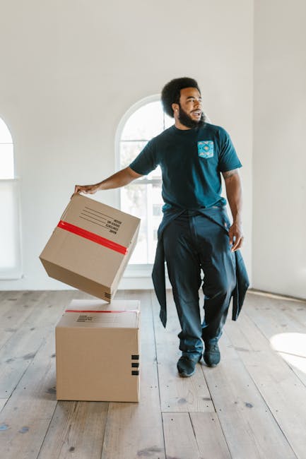 A man with dark curly hair and a beard is inside a bright, empty room with a large arched window, holding a cardboard box wrapped with red tape in his right hand while standing near two other cardboard boxes on the wooden floor. The boxes are standard packing cartons, some with visible labels and barcode stickers, likely prepared for a home relocation. The room has white walls and light natural lighting from the window, creating a clean, professional environment suitable for packing and moving activities. This image illustrates the process of furniture transport and packing during a household move, with the man acting as part of a removal service such as Man With a Van Leyton, focusing on last-minute or same-day removals in Leyton. The scene captures the essential elements of a professional moving company preparing items for transportation, possibly featuring blankets, trolleys, or other equipment not visible in the image.
