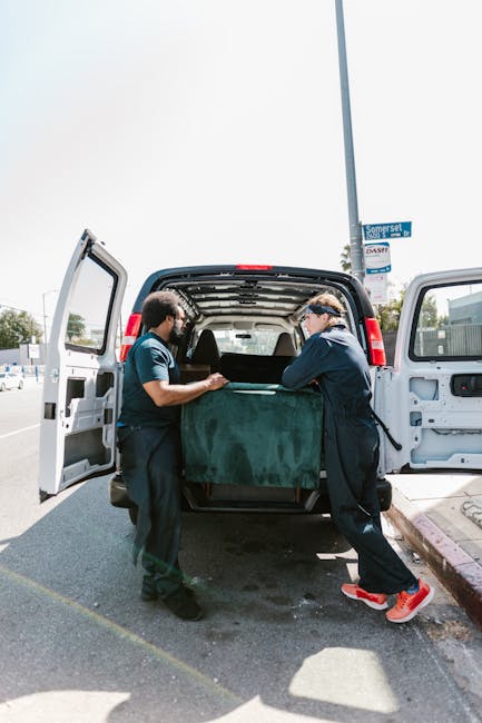 Two movers from Man With a Van Leyton are engaged in a home relocation process, standing outside a large white van with its rear doors open. They are carefully lifting or positioning a sizable, dark green upholstered piece of furniture, possibly a sofa or armchair, into the vehicle. The van is parked on the side of a street, adjacent to a pavement, with a lamppost visible to the right and a street sign that reads 'Somerset'. The interior of the van reveals a spacious cargo area, suitable for loading various household items. The environment is bright with natural daylight, indicating daytime, and the scene captures the careful handling involved in packing and moving furniture as part of a last-minute or same-day moving service, emphasizing the company's expertise in efficient and professional short-notice house removals.