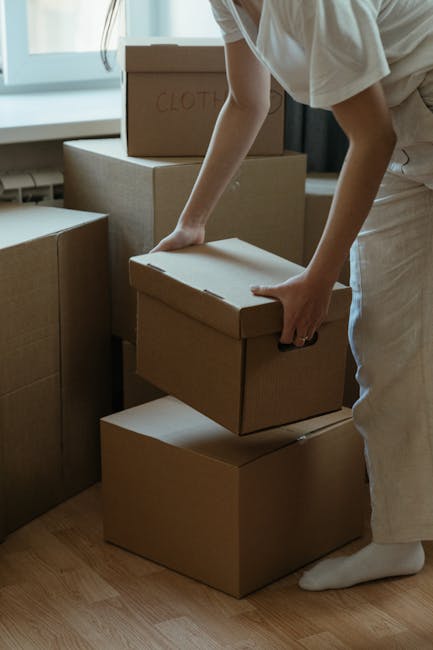 A man with dark curly hair and a beard is inside a bright, empty room with a large arched window, holding a cardboard box wrapped with red tape in his right hand while standing near two other cardboard boxes on the wooden floor. The boxes are standard packing cartons, some with visible labels and barcode stickers, likely prepared for a home relocation. The room has white walls and light natural lighting from the window, creating a clean, professional environment suitable for packing and moving activities. This image illustrates the process of furniture transport and packing during a household move, with the man acting as part of a removal service such as Man With a Van Leyton, focusing on last-minute or same-day removals in Leyton. The scene captures the essential elements of a professional moving company preparing items for transportation, possibly featuring blankets, trolleys, or other equipment not visible in the image.