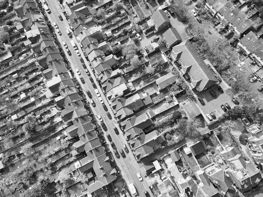 A black-and-white aerial photograph showing a densely packed residential street with rows of terraced houses on either side of a central road. The houses have pitched roofs, chimneys, and small front gardens or yards, with some featuring driveways or parking spaces. The central street is lined with parked cars along both curbs, and the road appears to be narrow, typical of urban housing areas in Leyton. Trees with bare branches are visible between the houses, indicating a possible winter season. The area around the houses includes narrow alleys, fences, and small garden spaces. This image captures the compact layout of the neighbourhood, relevant to house removals and moving logistics handled by Man With a Van Leyton, especially when planning home relocation or furniture transport within Waltham Forest.