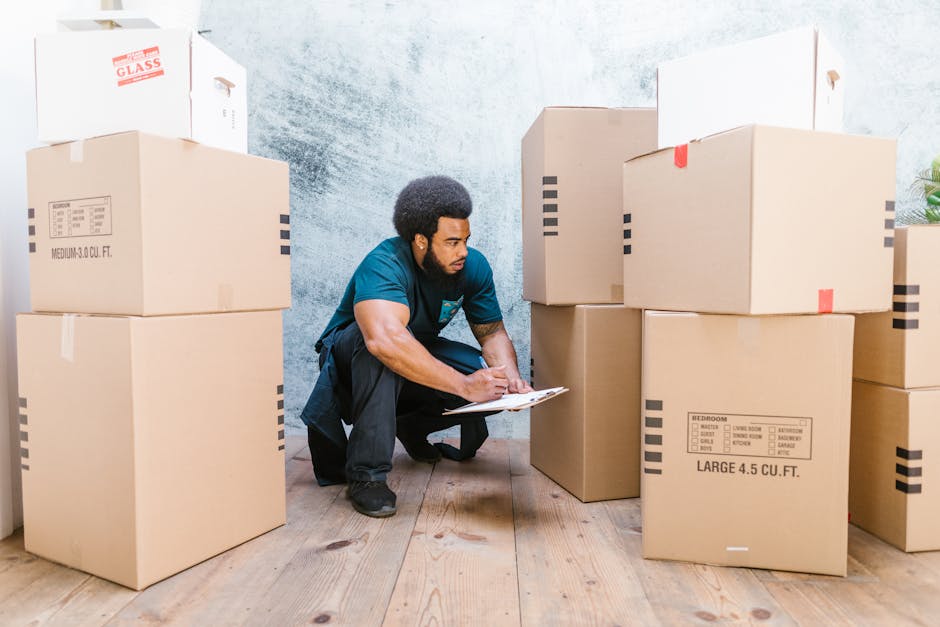 A man with a dark curly hairstyle and beard, dressed in a teal shirt and black pants, is crouching inside a room surrounded by several large cardboard boxes used for house removals. He is holding a clipboard and appears to be inspecting or making notes about the boxes, which are stacked on a wooden floor with some boxes positioned near a doorway and against a textured, light-colored wall. The boxes vary in size, some labeled with room designations such as 'bedroom' and 'large 4.5 cu.ft.', and are secured with clear or red tape. A few boxes are covered with protective plastic wrap, likely for safe furniture transport during packing and moving. In the background, there are additional boxes and a glimpse of a green plant, indicating a home environment prepared for a furniture transport or home relocation process. The scene reflects the careful planning and logistics involved in house removals, as managed by [COMPANY_NAME], emphasizing the importance of organized packing and the loading process in professional moving services.